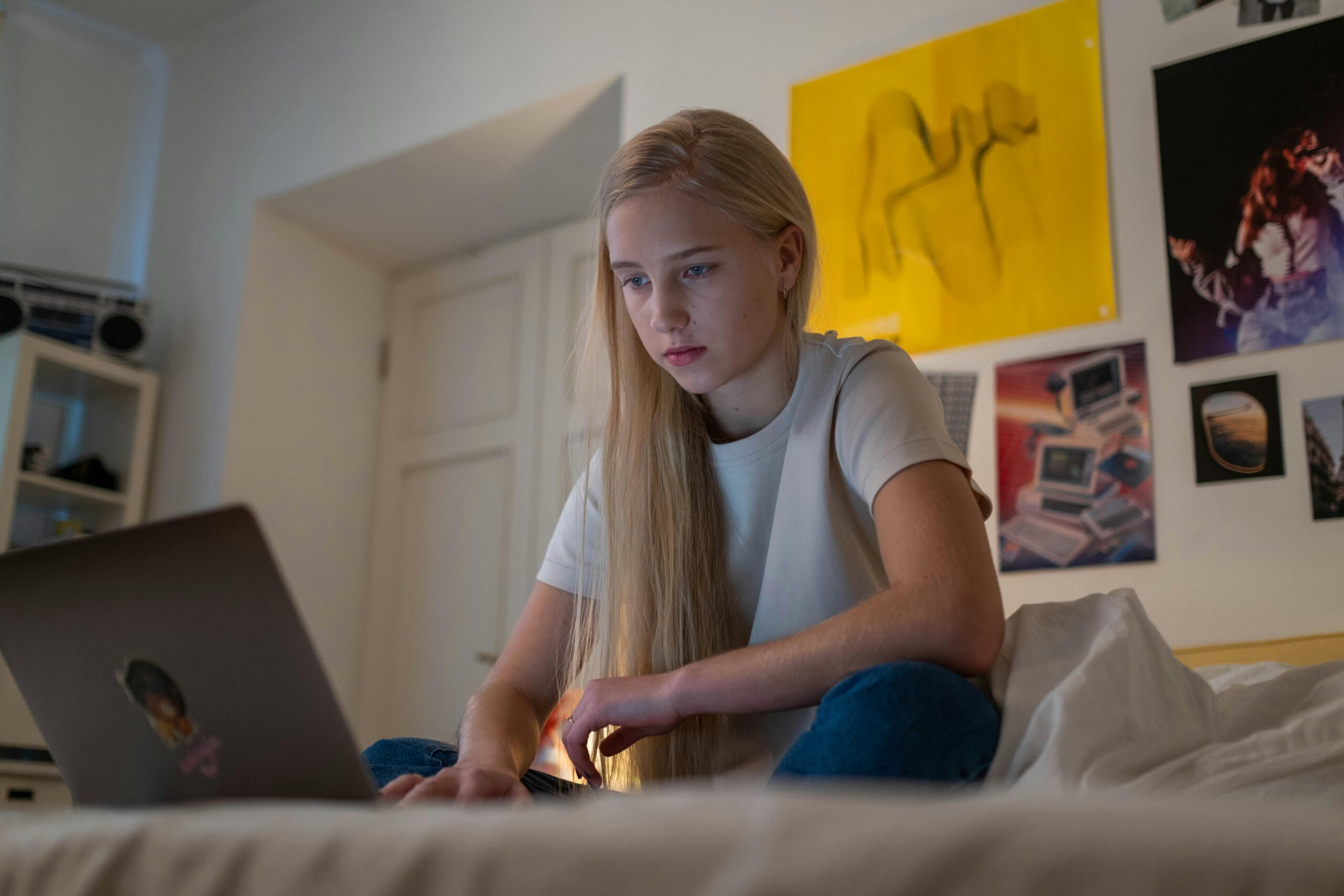 A young person with long blonde hair sits on a bed, using a laptop, and is focused on a session with a psychiatrist. The room has white walls decorated with colourful posters and photographs.